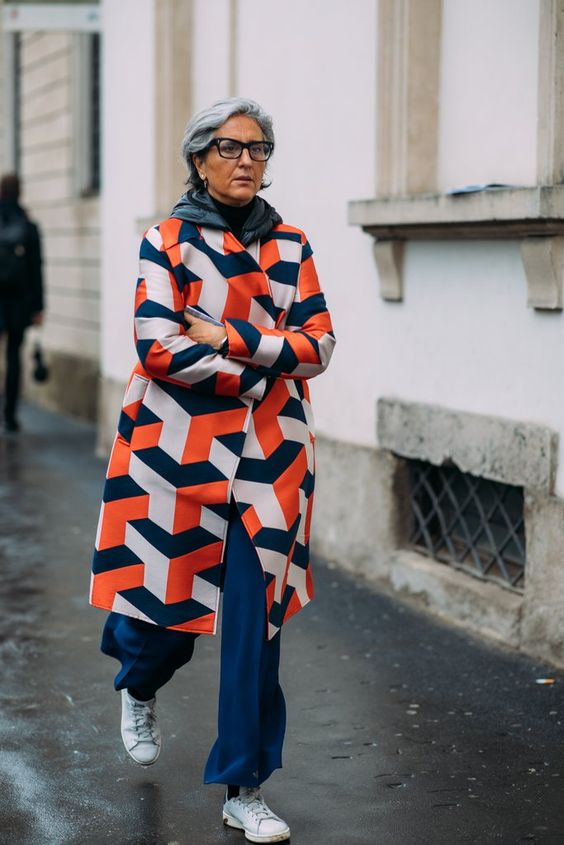 This woman is wearing a patterned coat. She has short grey hair and glasses.