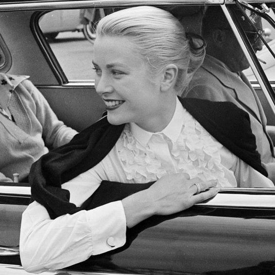 Grace Kelly is sitting in a car wearing a white ruffled shirt and a cartier love ring with her hair tied up